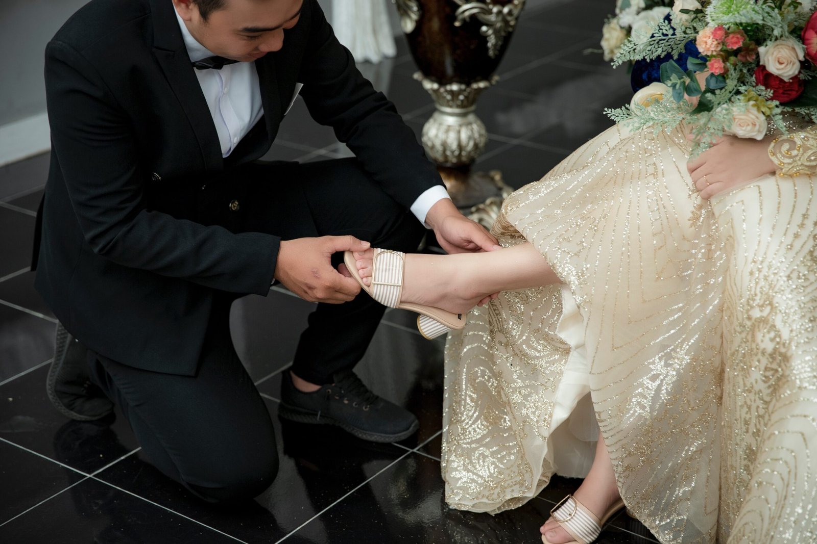 A couple on their wedding day with the groom showing he is an irresistible husband kneeling on one knee while helping his bride to fit her left shoe properly.