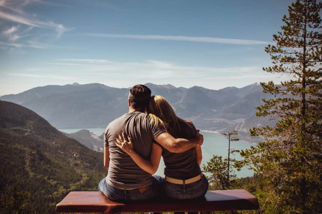 A couple cuddling each other while seated on a bench enjoying the views of picturesque mountain and ocean.