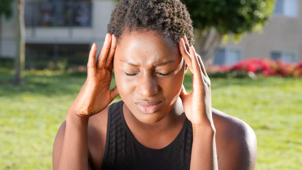 A Black lady holding her head and with her eyes closed in obvious pain from migraine headaches