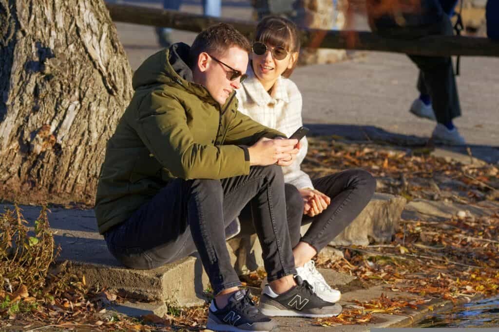 A couple is seated under a tree while the husband is reading out his text message from his smart phone while the wife pays attention to him.