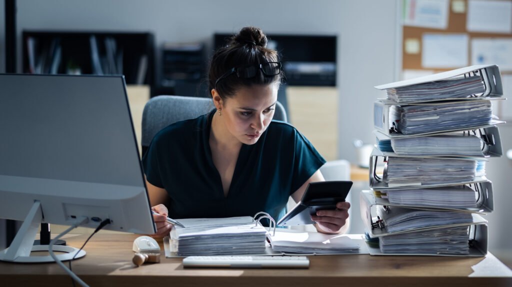 A female workaholic spouse who is working in her home office surrounded wby a huge pile of jacketed paper files