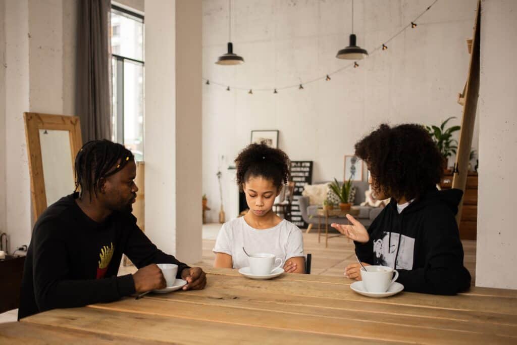 A Black family that are seated in their dining with cups of coffee and rebuking their young daughter whom is looking very sad.