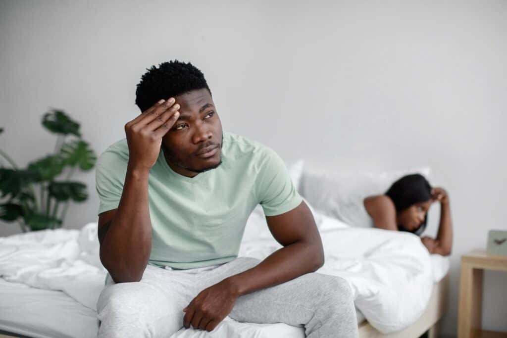 A Black couple who is engaged in marital conflict with the husband seating at the edge of the bed far away from his wife.