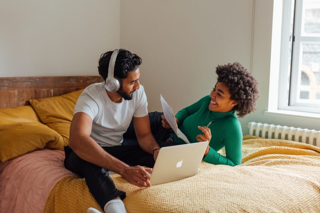 A Black couple who are seated on their bed chatting happily with the husband pausing from working on his laptop computer to give his wife his attention while she reads their financial report.