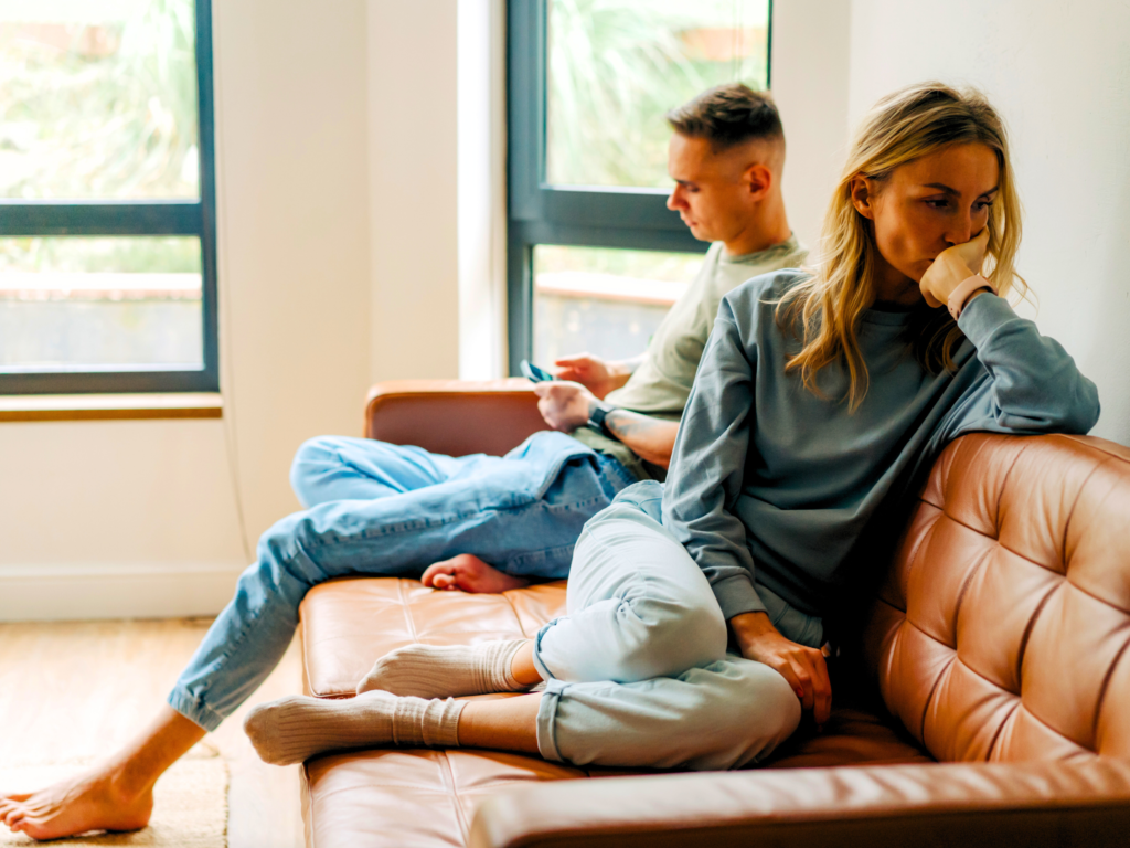 A sad wife looking away from her husband who is scrolling his phone as evidence of resentment in marriage.