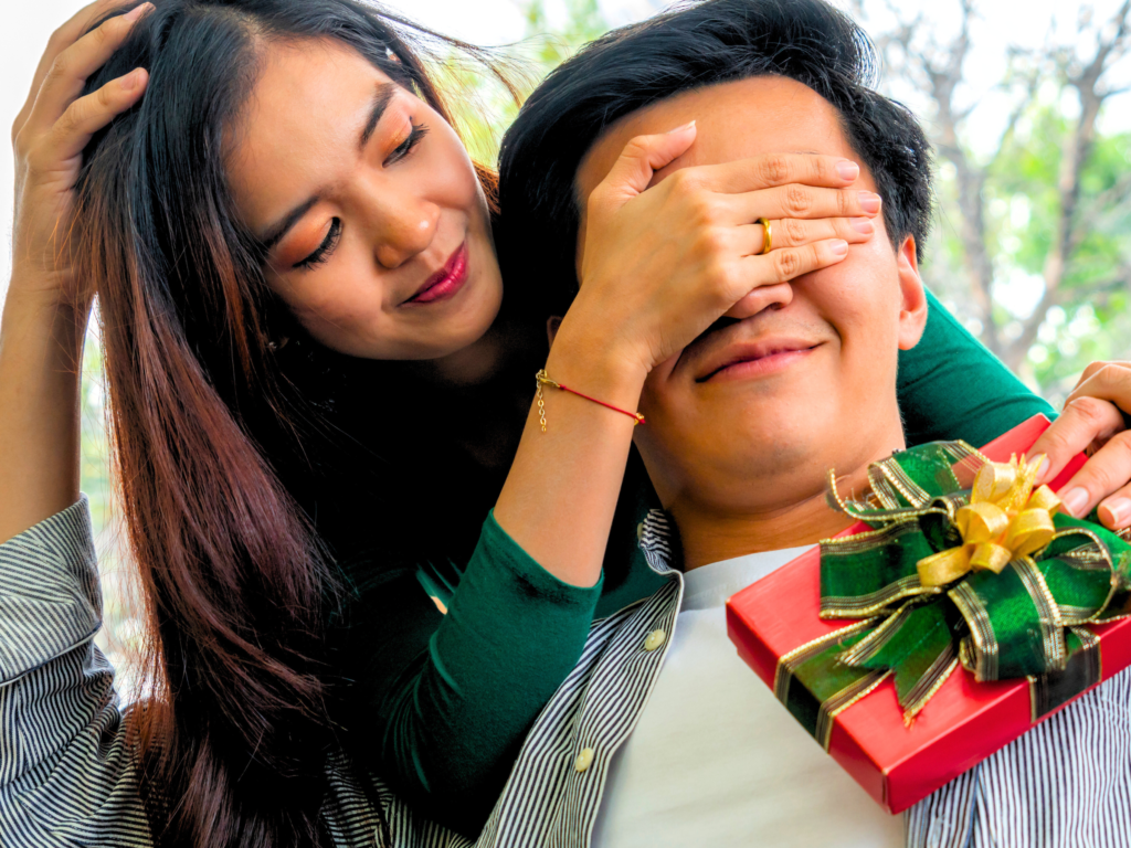 A wife surprising her husband with a wrapped gift to mark his birthday.